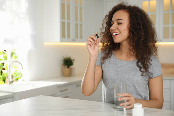 African-American woman with glass of water taking vitamin capsule in kitchen. Space for text - Foto, afbeelding