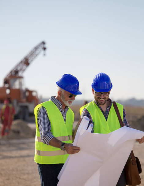 Two experienced engineers looking at blueprints and talking at building site with metal crane in background - Photo, Image