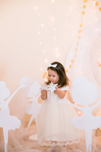 Cute little girl in a white princess dress with a bow on her hair plays with wooden ballerinas among the golden stars on the background of a large white wooden moon and garlands. Children's decor. Children holiday - Foto, Bild