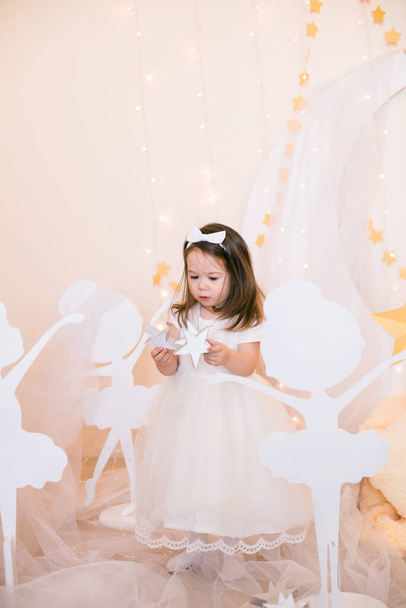 Cute little girl in a white princess dress with a bow on her hair plays with wooden ballerinas among the golden stars on the background of a large white wooden moon and garlands. Children's decor. Children holiday - Foto, Imagen