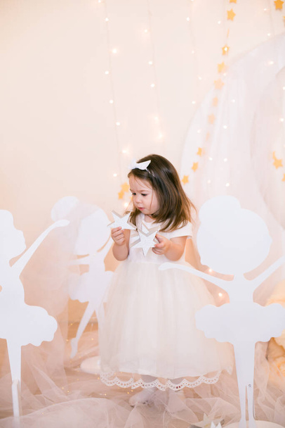 Cute little girl in a white princess dress with a bow on her hair plays with wooden ballerinas among the golden stars on the background of a large white wooden moon and garlands. Children's decor. Children holiday - Photo, Image