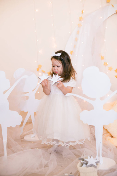 Cute little girl in a white princess dress with a bow on her hair plays with wooden ballerinas among the golden stars on the background of a large white wooden moon and garlands. Children's decor. Children holiday - Fotografie, Obrázek