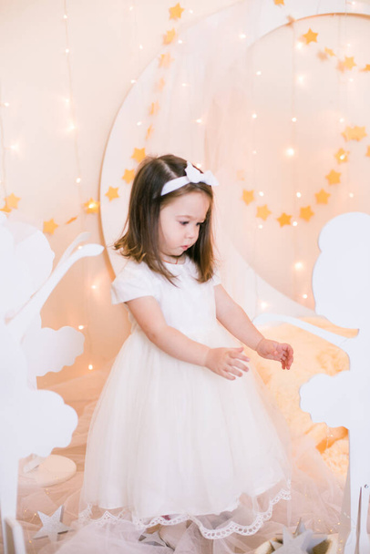 Cute little girl in a white princess dress with a bow on her hair plays with wooden ballerinas among the golden stars on the background of a large white wooden moon and garlands. Children's decor. Children holiday - Фото, зображення