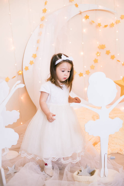 Cute little girl in a white princess dress with a bow on her hair plays with wooden ballerinas among the golden stars on the background of a large white wooden moon and garlands. Children's decor. Children holiday - Фото, зображення