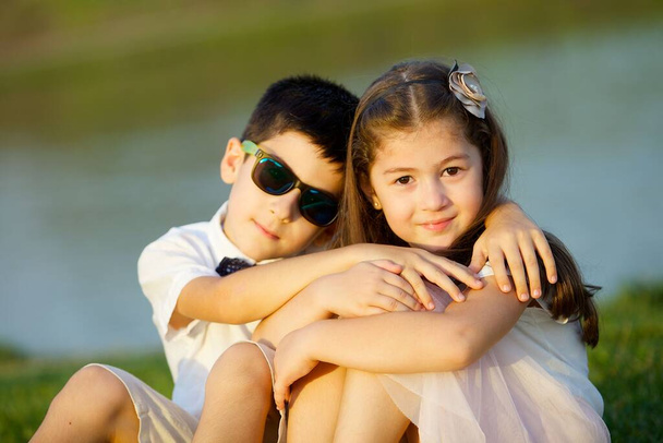 Ένα αγόρι και ένα κορίτσι κάθονται στο βάθος μιας λίμνης ή ενός ποταμού. Φύση και καλοκαιρινές διακοπές. - Φωτογραφία, εικόνα