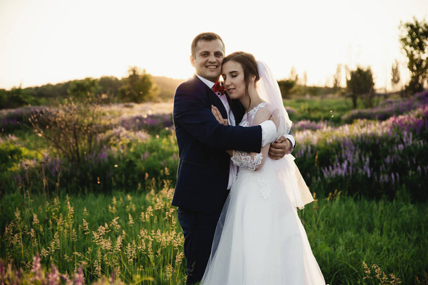 Bride and groom hugging at sunset.Happy bride and groom smiling.Happy bride and groom after the wedding ceremony.Wedding day.Beautiful young couple in a field with flowers,wedding day in the lives of young people - Fotó, kép