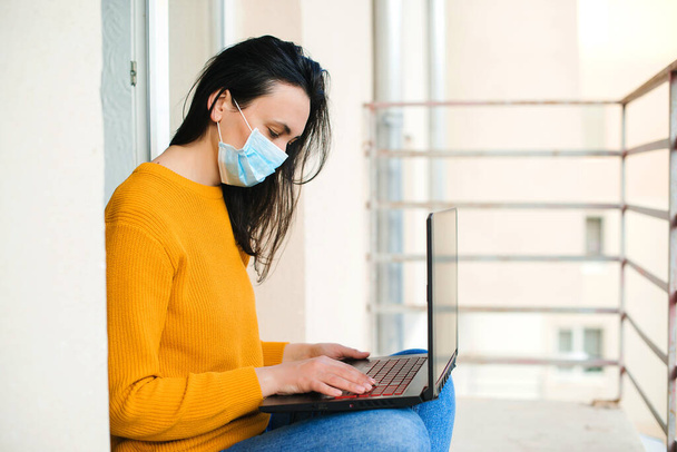 Nő maszkot visel. Egy lány dolgozik egy laptopon az erkélyen. Technológia, szabadidő és életmód. Szabadúszó, távoli munka. Coronavirus karantén. Otthonról dolgozom. Távoktatás a karantén ideje alatt. - Fotó, kép