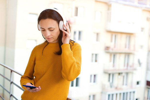 Young woman with headphones breathing fresh air on a balcony. Woman using mobile phone and wireless headphones. Girl listen music at balcony. Stay home. Self isolation at home. Coronavirus quarantine. - Photo, Image