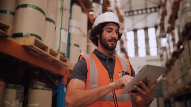 背景に商品のスタックを持つ大規模な倉庫でデジタルタブレットを使用して白いハード帽子の若い男を笑顔 - 映像、動画