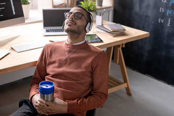 Young restful programmer in headphones holding mug with drink while sitting on chair against his workplace at break - Foto, Imagen