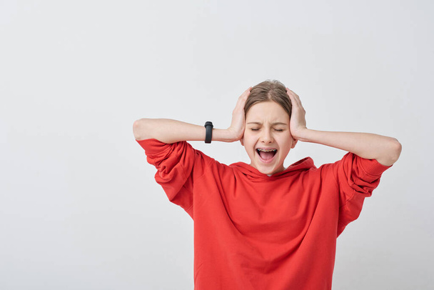 Шокированная или истеричная девочка-подросток в красной толстовке закрывает уши, прикасаясь к голове и крича на белом фоне
 - Фото, изображение