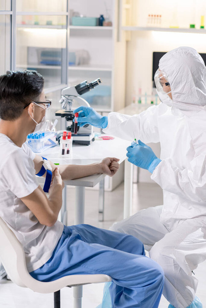Infizierter Chinese mit Maske und Laborarbeiter in Schutzanzügen, Handschuhen und Brille am Schreibtisch im medizinischen Labor - Foto, Bild