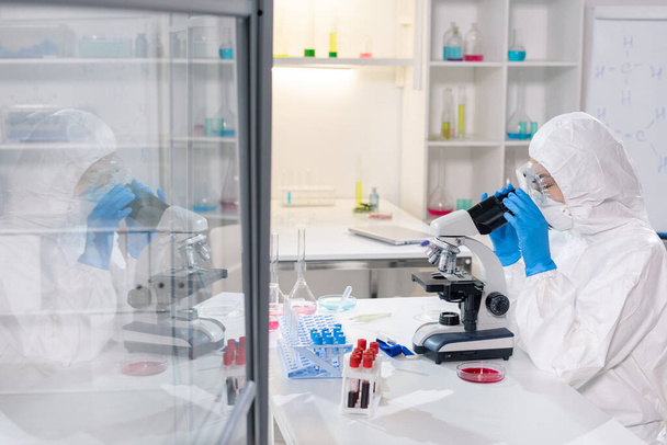 Σύγχρονη νεαρός επιστήμονας ή ερευνητής σε προστατευτικές φόρμες, μάσκα και γάντια κοιτάζοντας στο μικροσκόπιο, ενώ τη μελέτη του νέου ιού - Φωτογραφία, εικόνα