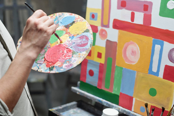 Kéz a férfi művész festőecsettel és paletta keverés színek, miközben áll a vászon előtt befejezetlen festmény - Fotó, kép