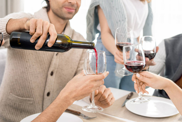 Junger Mann schenkt Rotwein aus Flasche in Weingläser seiner Freunde ein, während er gemeinsam den Urlaub am servierten Tisch feiert - Foto, Bild