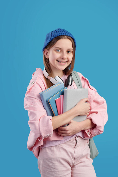 Całkiem sprytna nastolatka z książkami przy klatce piersiowej stojąca przed kamerą na niebieskim tle i patrząca na ciebie z uśmiechem - Zdjęcie, obraz
