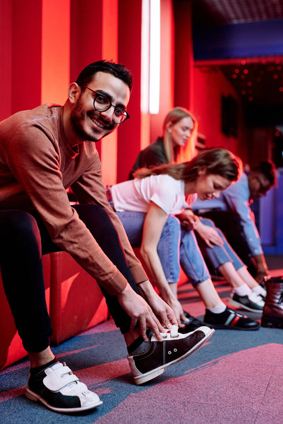 Glücklicher junger Mann mit gemischter Rasse in Freizeitkleidung und seine Freunde im Hintergrund bereiten sich darauf vor, Bowling zu spielen, während sie die Schuhe wechseln - Foto, Bild