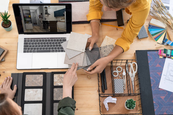 Dos jóvenes diseñadoras de interiores discuten muestras de paneles por lugar de trabajo mientras eligen uno para las paredes de la habitación en la pantalla del ordenador portátil
 - Foto, Imagen