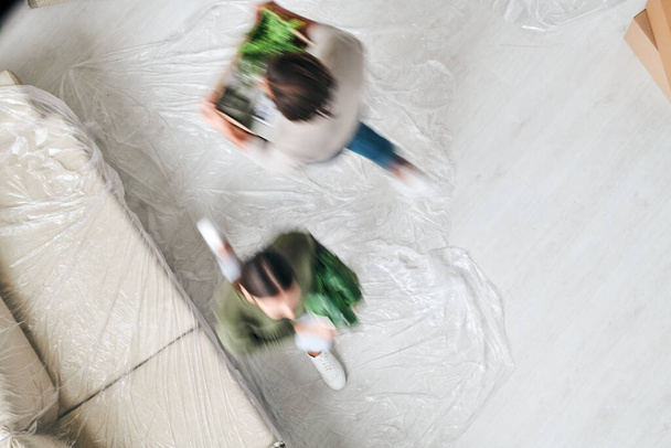 Вид сверху на размытые муж и жена, несущие зеленые домашние растения, двигаясь по дивану в гостиной новой квартиры
 - Фото, изображение