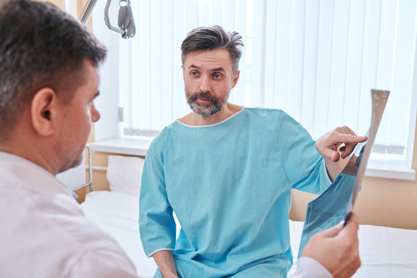 Maturo paziente barbuto in abito da ospedale seduto sul letto e che indica l'immagine a raggi X mentre chiede al medico circa la sua malattia
 - Foto, immagini
