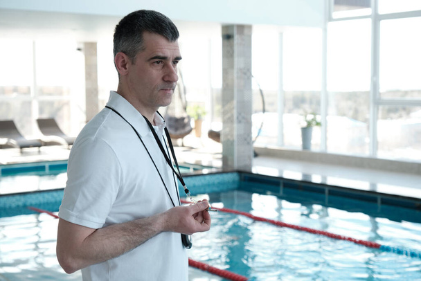 Grave allenatore di nuoto maturo in piedi a piscina e utilizzando cronometro durante l'allenamento nuotatore
 - Foto, immagini