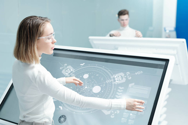 Молодая женщина в очках работает с цифровой схемой на интерактивном дисплее в современном офисе
 - Фото, изображение