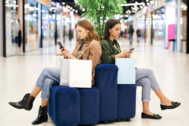 Alışveriş merkezinin lobisinde puf puf üzerinde oturan ve alışveriş için özel teklifler ararken akıllı telefon kullanan ciddi genç kadınlar. - Fotoğraf, Görsel