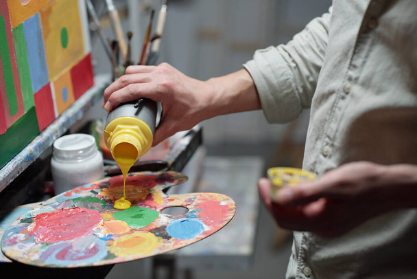 Hand of young painter adding yellow color on palette before start painting while standing in front of easel in studio of arts - Photo, Image