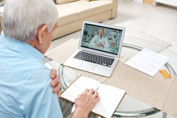 Over shoulder view of elderly white-haired man sitting at table and using laptop while asking doctor about pain online - Photo, Image