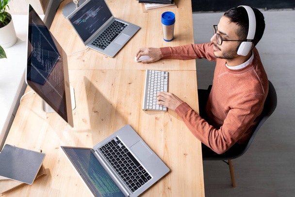 Занятый молодой разработчик программного обеспечения в наушниках сидит за столом перед двумя ноутбуками и монитором компьютера во время работы
 - Фото, изображение