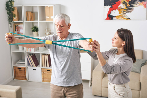 Привлекательная молодая женщина личный тренер объясняя упражнения с резиной для пожилого человека, чтобы укрепить его мышцы позвоночника
 - Фото, изображение
