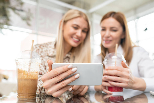 Hand of young female shopper with smartphone showing her friend online goods or clothes while both resting in cafe and having drinks - Foto, imagen