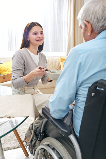 Smiling pretty nurse feeding senior disabled man in wheelchair and talking to him in living room - Photo, image