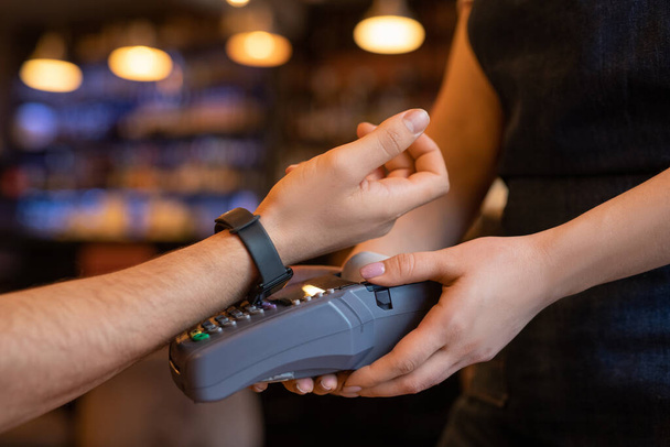 Χέρι του νεαρού πελάτη του καφέ ή εστιατόριο κρατώντας βραχίονα με SmartWatch πάνω μηχάνημα πληρωμής, ενώ πληρώνουν για ποτό ή φαγητό - Φωτογραφία, εικόνα