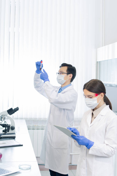 Mladá žena v ochranném pracovním oděvu rolování v tabletu, zatímco její kolegyně při pohledu na baňku s novou vakcínou nebo chemickou látkou v laboratoři - Fotografie, Obrázek