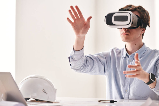 Giovane ingegnere concentrato seduto a tavola e mani contorte mentre utilizza il dispositivo di realtà virtuale per realizzare un progetto di costruzione 3D
 - Foto, immagini