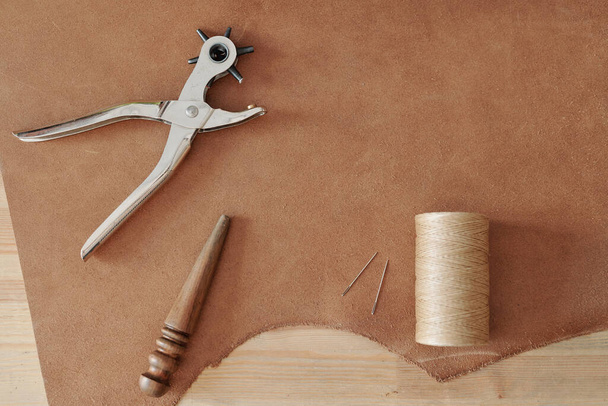 Overview of handtool for making holes, spool with light threads, two needles and wooden tool on part of beige suede - Zdjęcie, obraz