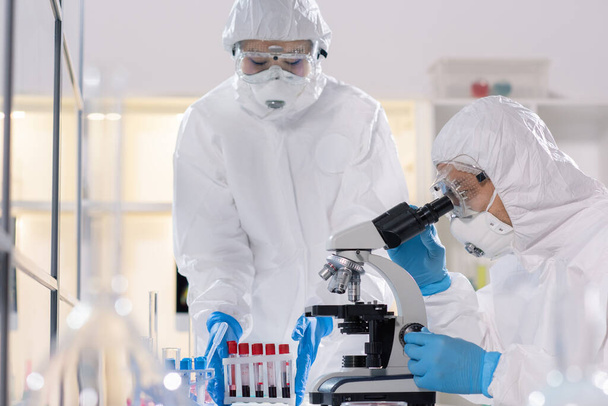Científico joven en ropa de trabajo protectora mirando en el microscopio mientras estudia el nuevo virus con su colega en el laboratorio médico
 - Foto, imagen