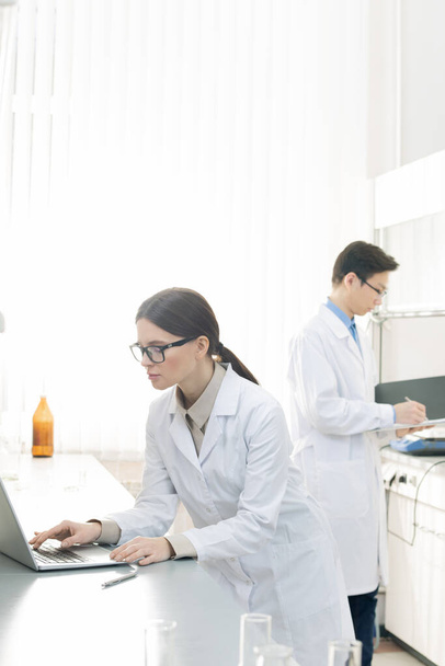 Досить молода жінка-працівник лабораторії у білому костюмі та окулярах стоїть за столом перед дисплеєм ноутбука, вивчаючи нові можливості вірусу
 - Фото, зображення