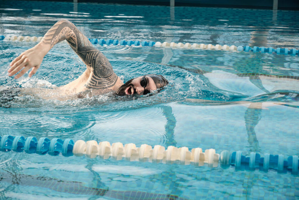 Atletische jonge man met baard in bril en pet zwemmen beroerte in het zwembad tijdens de training voor de wedstrijd - Foto, afbeelding