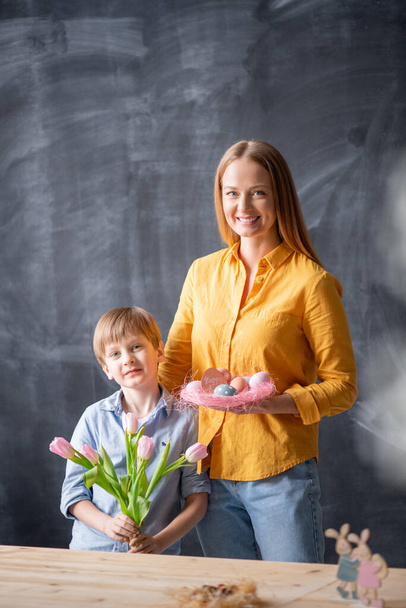 Portrét šťastné mladé matky se synem stojící s tulipány a velikonočním hnízdem proti tabuli - Fotografie, Obrázek
