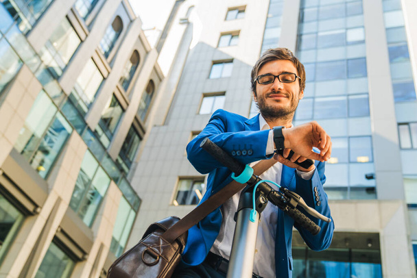 Jeune homme d'affaires debout avec scooter électrique et regardant l'heure sur la montre tout en se tenant dans la rue de la ville
 - Photo, image