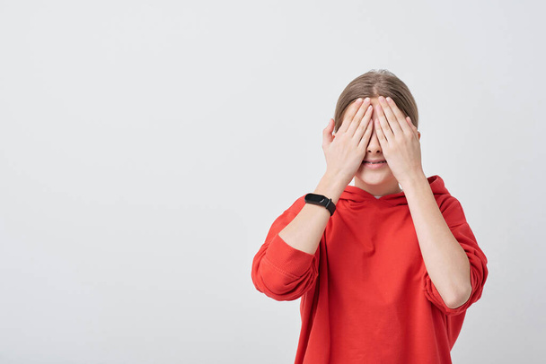 Usmívající se dospívající dívka v červené mikině zakrývající si oči rukama během schovávat-a-hledat hru, zatímco stojí před kamerou - Fotografie, Obrázek