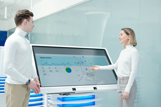 Positive friendly young woman pointing at interactive display and analyzing statistics of product distribution with colleague - Photo, Image
