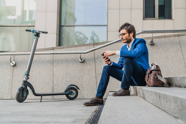 Side view of young businessman sitting on stairs using phone near electric scooter - Zdjęcie, obraz
