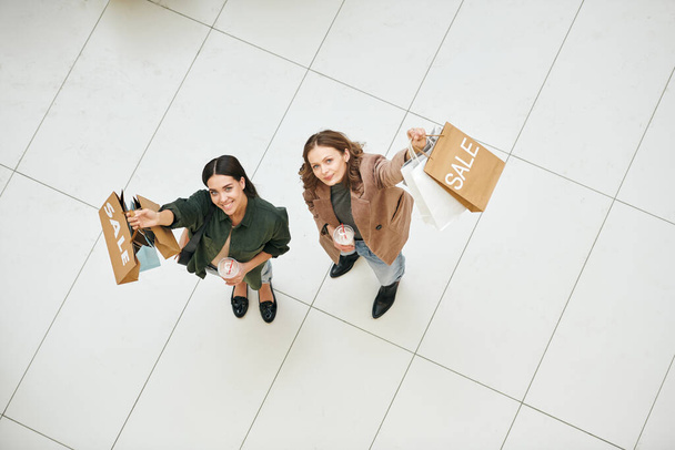 Vista superior de las mujeres positivas agitando bolsas de compras y mirando a la cámara en el vestíbulo del centro comercial
 - Foto, imagen
