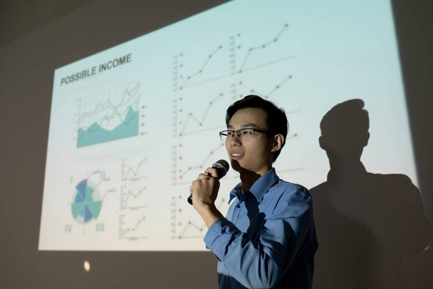 投資家に自分のビジネスアイデアを提示しながら、投影画面に立ってマイクに話す眼鏡の若いアジアの男を確信 - 写真・画像