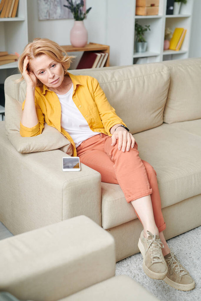 Ältere selbstisolierte Geschäftsfrau in Freizeitkleidung sitzt auf der Couch und wartet auf einen Anruf von Freund oder Kollege, während sie zu Hause bleibt - Foto, Bild
