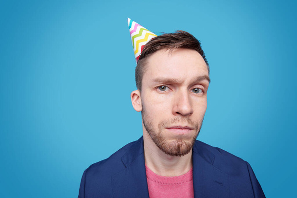 Suspicious young man in jacket, pullover and birthday cap looking at you in isolation against blue background - Photo, Image