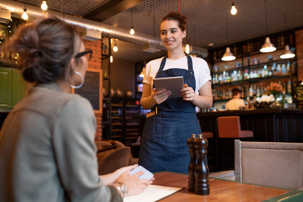 Щаслива молода офіціантка з цифровим планшетом стоїть на одному зі столів перед жіночим гостем і приймає її замовлення в кафе або ресторані
 - Фото, зображення